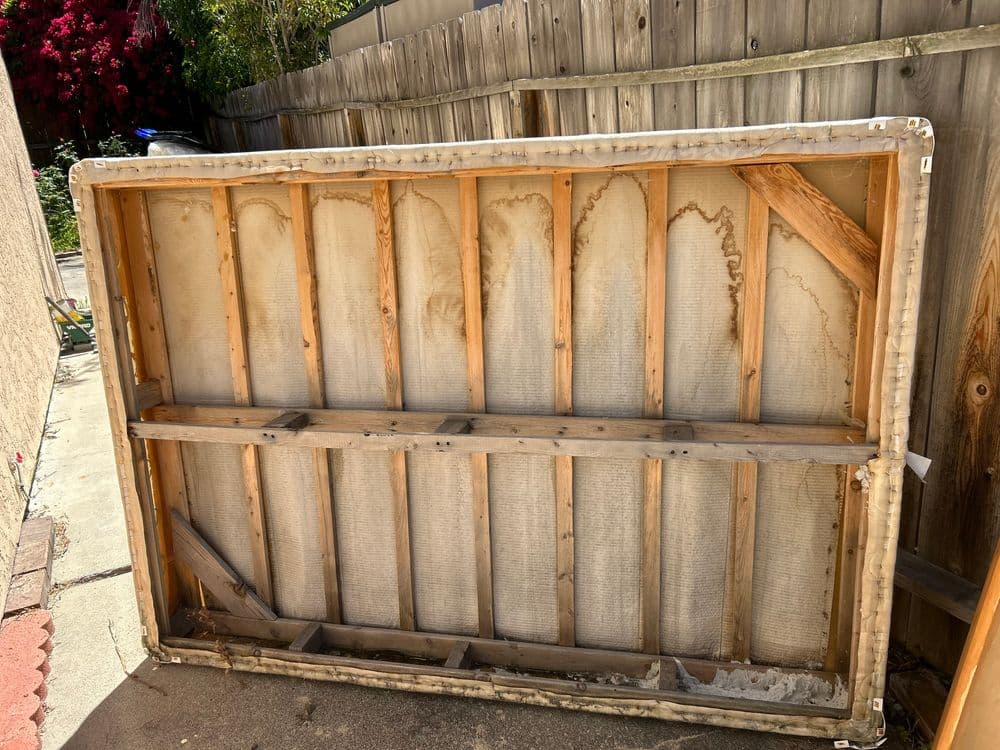 Old, damaged mattress frame leaning against a wooden fence in a backyard.