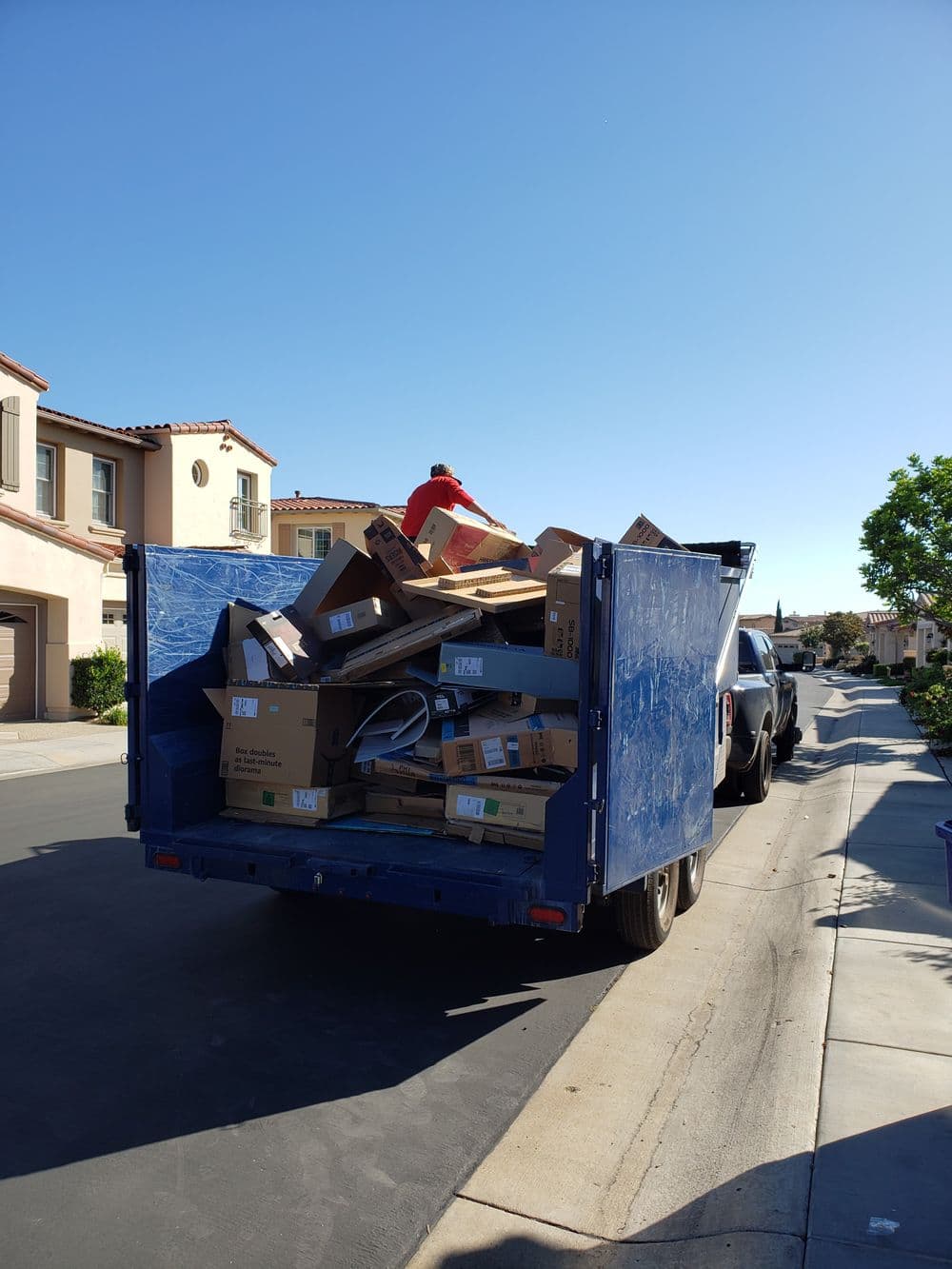 Worker loading cardboard boxes into a blue waste truck on a residential street.