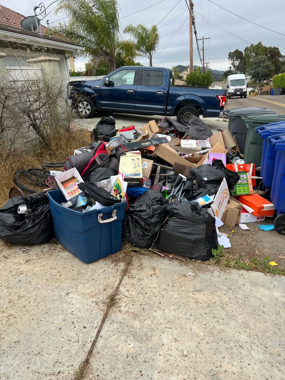 Piled trash and recycling bins next to a parked blue truck on a residential street.