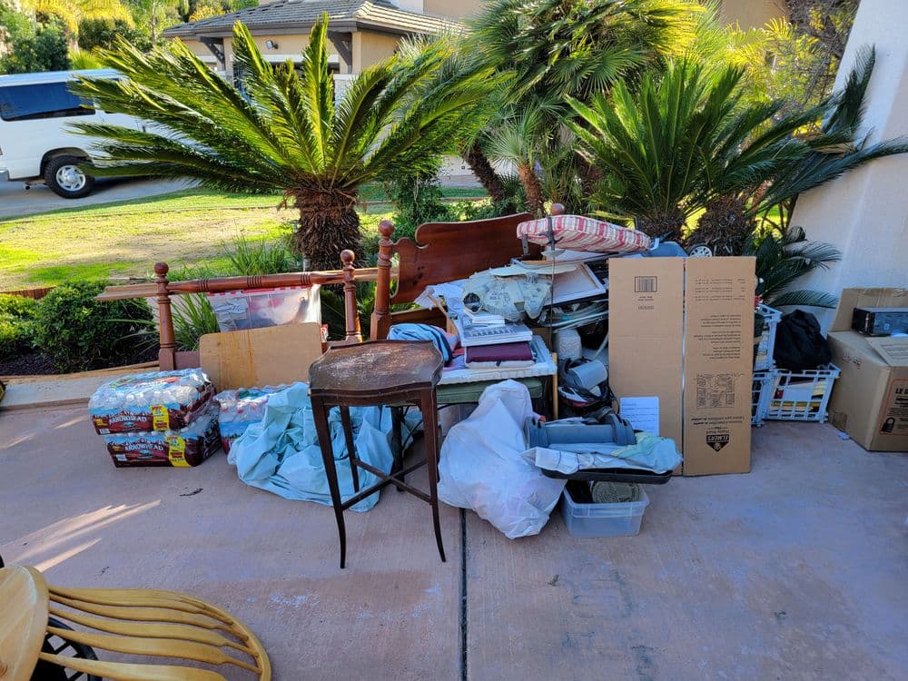 Piled clutter including boxes, furniture, and household items on a driveway surrounded by palm trees.