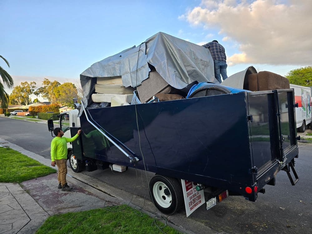 Moving truck loaded with furniture, a worker securing items under a tarp on a residential street.