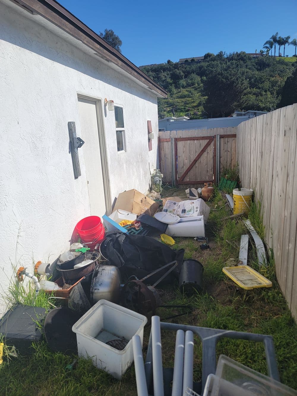 Cluttered outdoor area beside a house with various discarded items and gardening supplies.