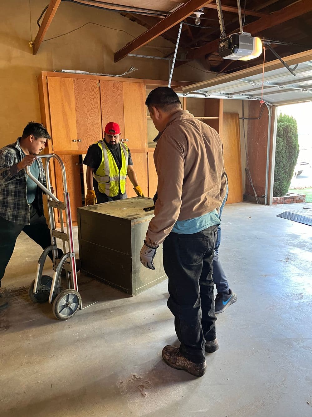 Three men moving a large box in a garage, with one using a hand truck.