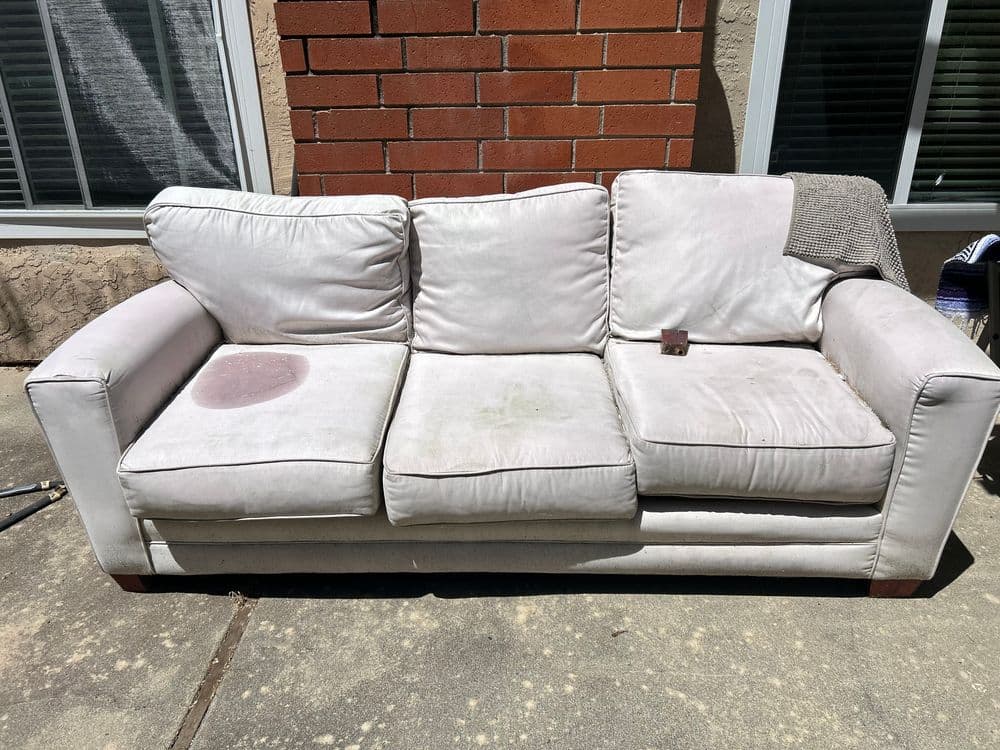 Worn white leather sofa with stains and a textured throw, set against a brick wall.