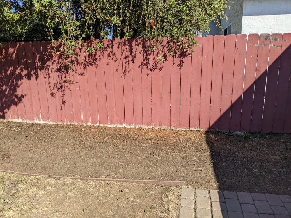 Red wooden fence with overhanging greenery and bare ground alongside a paved path.