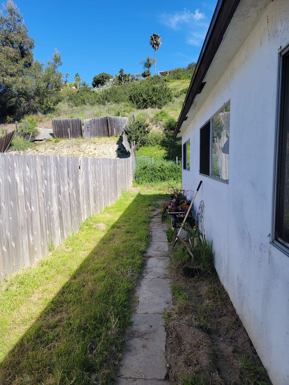 Pathway alongside a white house, bordered by a wooden fence and greenery under a blue sky.