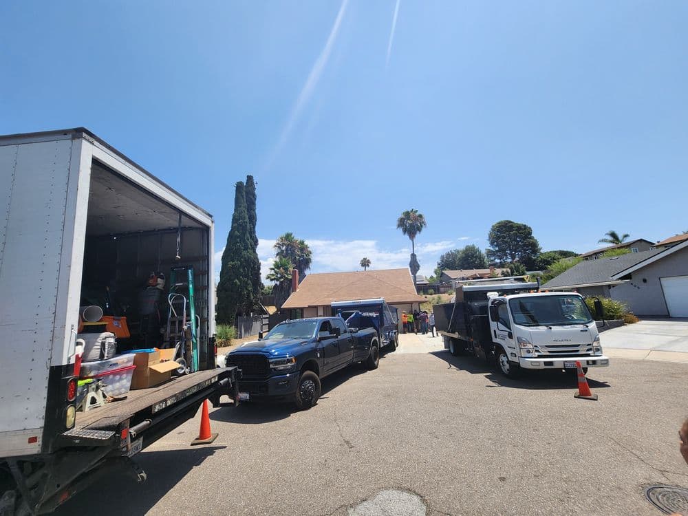 Two moving trucks parked in a residential area with palm trees and a house in the background.