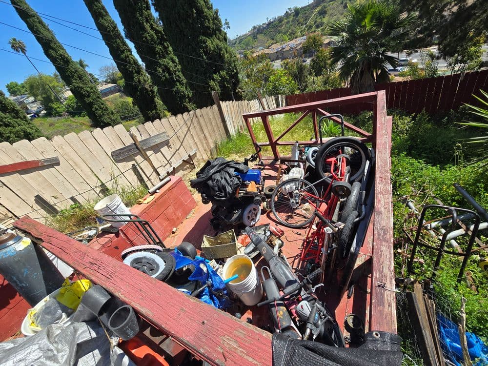 Cluttered backyard trailer filled with tools, machinery, and vegetation against a wooden fence.