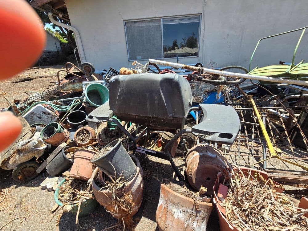 Piled-up junk including pots, tools, and a grill near a house in a cluttered yard.