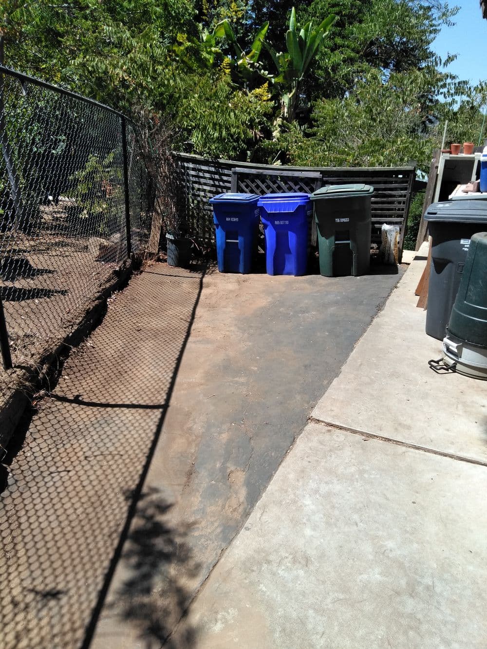 Recycling and waste bins lined along a concrete pathway in a backyard garden.