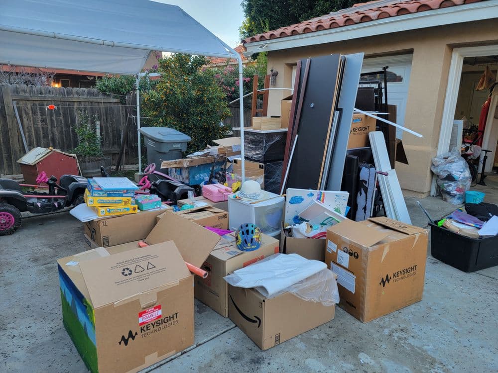 Cluttered garage filled with cardboard boxes and miscellaneous items under a canopy.