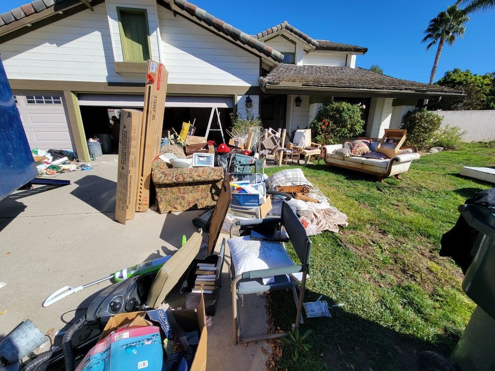 Cluttered front yard of a house with piles of furniture and debris outside a garage.