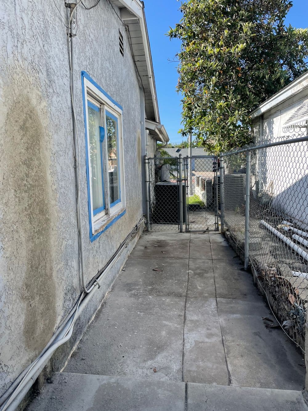 Narrow concrete pathway beside a house leading to a fenced area with a tree in background.