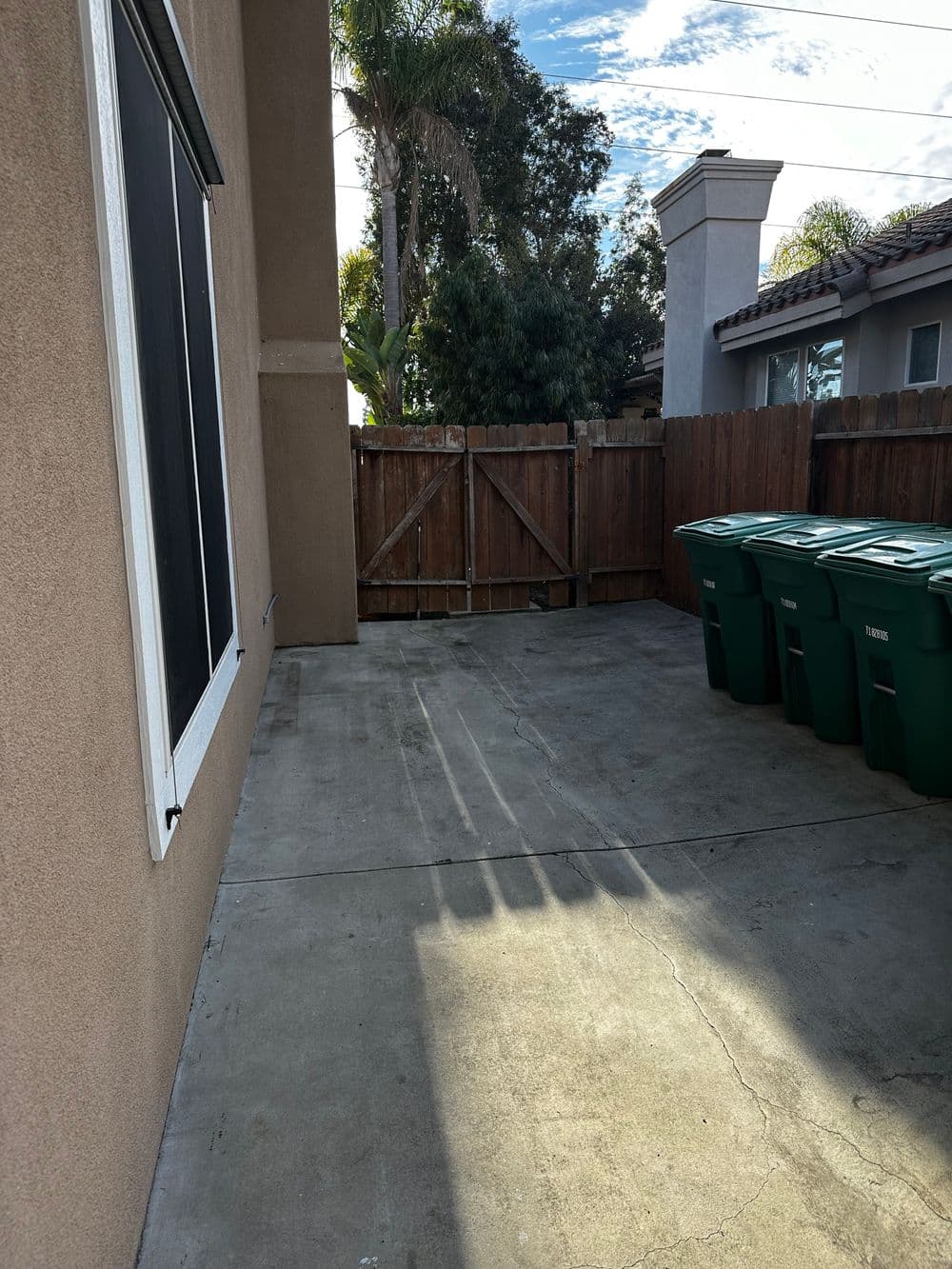 Patio area with concrete floor, green trash bins, and wooden fence, under blue sky with clouds.